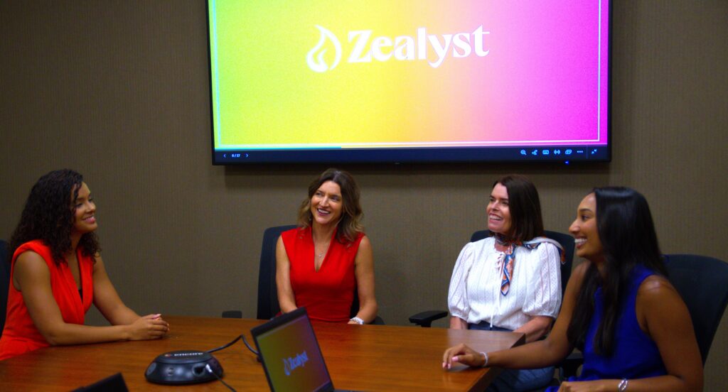 Four smiling women at a conference table with the Zealyst logo on a screen behind them
