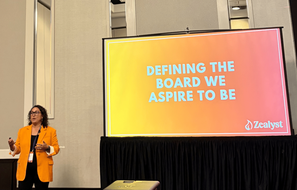 Woman speaking in front of a screen displaying "DEFINING THE BOARD WE ASPIRE TO BE" and the Zealyst logo