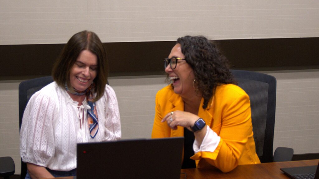 Two women laughing and smiling, one in a white blouse, the other in a bright yellow jacket