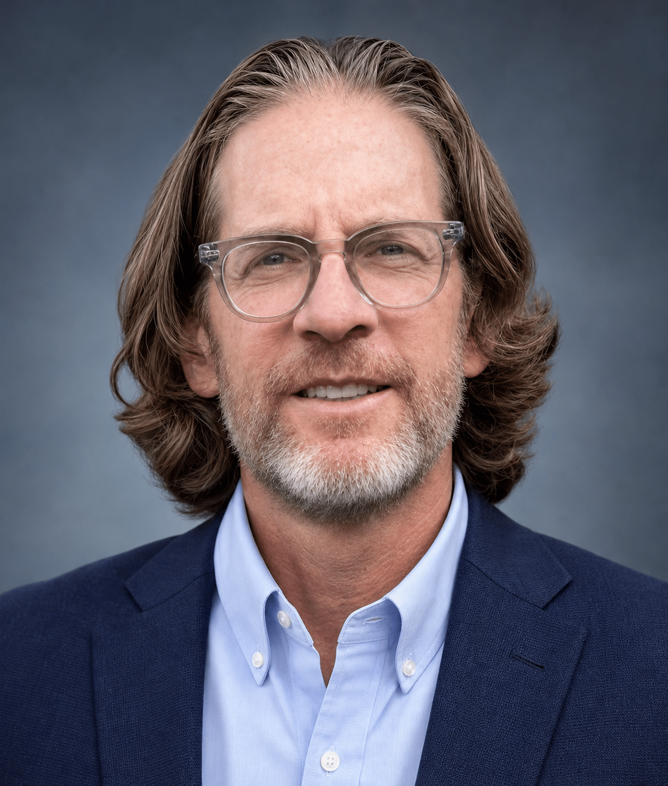 Man with brown hair, beard, glasses, light blue shirt, and dark blue blazer smiling at the camera