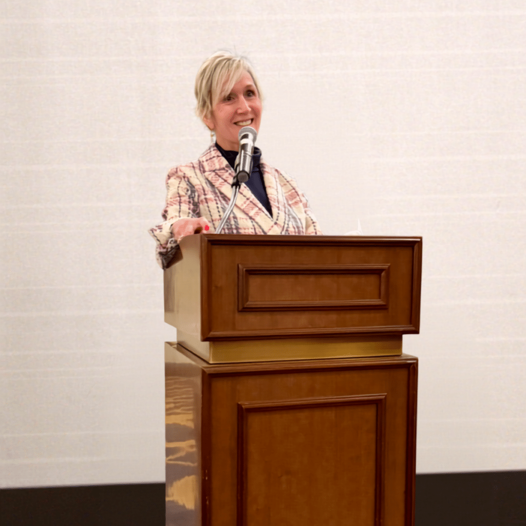 Woman with blonde hair smiling while speaking into a microphone at a wooden podium